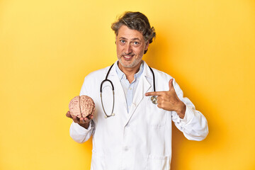 Doctor holding a brain model, yellow studio backdrop person pointing by hand to a shirt copy space, proud and confident