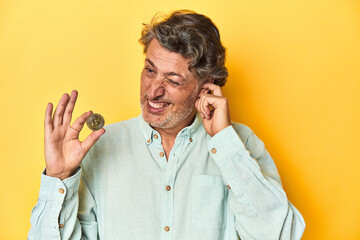 Man holding a physical bitcoin, yellow backdrop covering ears with hands.