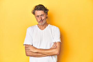Middle-aged man posing on a yellow backdrop frowning face in displeasure, keeps arms folded. - obrazy, fototapety, plakaty
