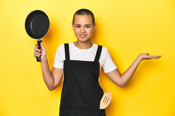 Cook woman holding a pan, yellow studio background showing a copy space on a palm and holding another hand on waist.