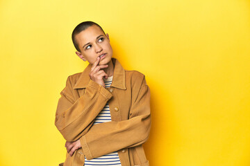 Woman in striped tee and denim shirt, yellow backdrop looking sideways with doubtful and skeptical expression.