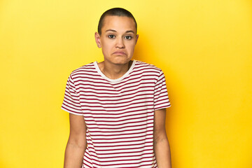Shaved head woman in red striped tee, yellow backdrop shrugs shoulders and open eyes confused.