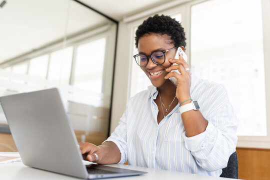 Confident young entrepreneur using laptop, talking on mobile phone in contemporary co-working space. Stylish african-american woman with short haircut sitting at desk, has pleasant phone conversation