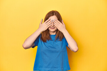 Redhead in blue t-shirt on yellow backdrop afraid covering eyes with hands.