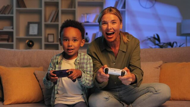 Blonde mom and little multiracial son are sitting on the sofa at home in the evening and playing a video game. Relatives enjoy spending time together, playing races on gamepads.