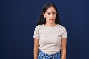 Young hispanic woman standing over blue background skeptic and nervous, frowning upset because of...