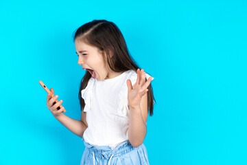 Angry Caucasian kid girl wearing  white t-shirt over blue background screaming on the phone, having...