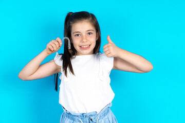 Caucasian kid girl wearing  white t-shirt over blue background holding an invisible braces aligner and rising thumb up, recommending this new treatment. Dental healthcare concept.