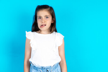 Portrait of dissatisfied Caucasian kid girl wearing  white t-shirt over blue background  smirks face, purses lips and looks with annoyance at camera, discontent hearing something unpleasant