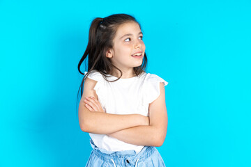 Caucasian kid girl wearing  white t-shirt over blue background arms crossed look empty space...
