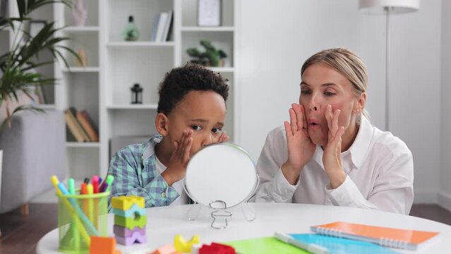Cute little multiracial boy with speech problems practicing with occupational therapist, practicing and looking at mirror in office. The concept of childhood autism.