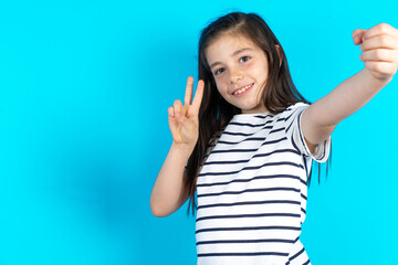 Fototapeta premium Positive Caucasian kid girl wearing striped T-shirt over blue background take selfie make v-sign