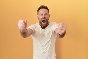 Middle age man with beard standing over yellow background angry and mad raising fists frustrated and furious while shouting with anger. rage and aggressive concept.