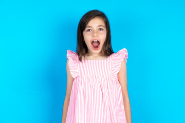 Oh my God. Surprised caucaisna kid girl wearing pink dress over blue background stares at camera...