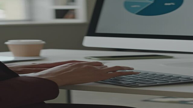 Vertical shot of unrecognizable female hands typing on keyboard while analyzing financial data on diagram on computer screen at white work desk in office