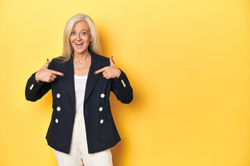 Middle-aged Caucasian woman in business jacket, yellow studio surprised pointing with finger, smiling broadly.