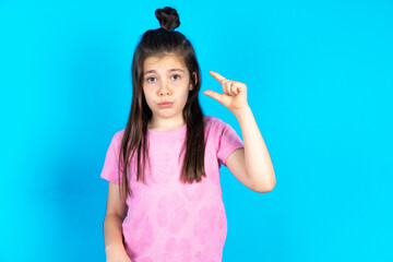 Caucasian kid girl wearing pink t-shirt over blue background purses lip and gestures with hand,...