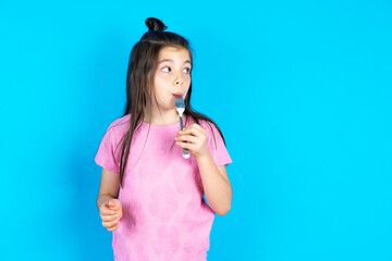 Photo of dreamy Caucasian kid girl wearing pink t-shirt over blue background lick fork look empty...