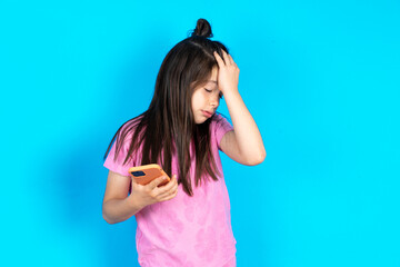 Upset depressed Caucasian kid girl wearing pink t-shirt over blue background makes face palm as...