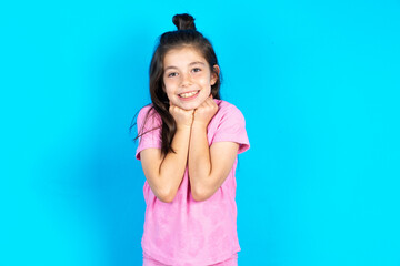 Obraz na płótnie Canvas Satisfied Caucasian kid girl wearing pink t-shirt over blue background touches chin with both hands, smiles pleasantly, rejoices good day with lover