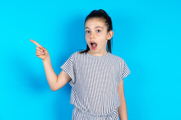 Emotive Caucasian kid girl wearing striped dress over blue background keeps jaw dropped from shock demonstrates amazing promo points right on blank space demonstrates big shopping sale. Advertisement