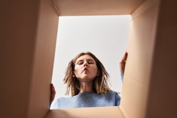 Beautiful woman opening cardboard box making fish face with mouth and squinting eyes, crazy and...