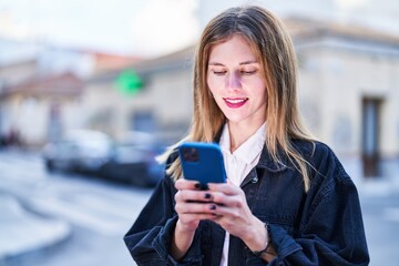 Young blonde woman using smartphone smiling at street
