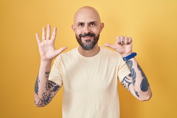Hispanic man with tattoos standing over yellow background showing and pointing up with fingers number six while smiling confident and happy.