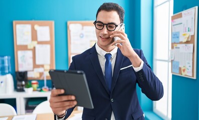 Young hispanic man business worker talkiong on smartphone using touchpad at office