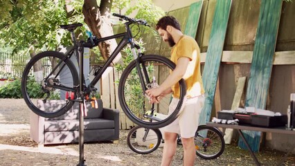 Healthy committed male cyclist carefully loosening and dismantling damaged tire from bicycle body. Sporty young man cautiously unfastening bicyle wheel and securing front fork in home yard.