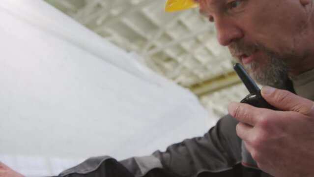Medium close up shot of mature worker in hardhat using walkie talkie and touching surface of white marble slab at stone factory