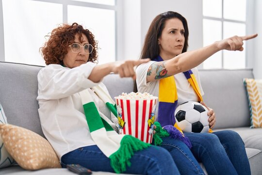 Hispanic mother and daughter watching football supporting team pointing with finger to the camera and to you, confident gesture looking serious - Powered by Adobe