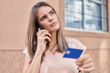 Young beautiful hispanic woman talking on smartphone using credit card with doubt expression at street