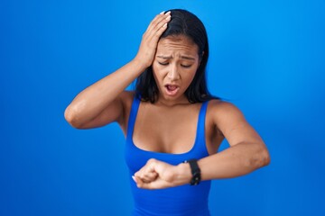 Hispanic woman standing over blue background looking at the watch time worried, afraid of getting...