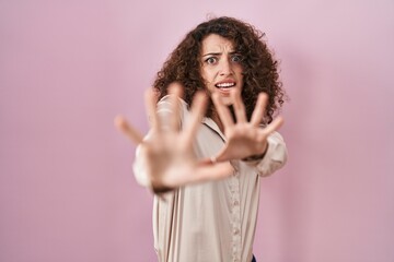Hispanic woman with curly hair standing over pink background afraid and terrified with fear expression stop gesture with hands, shouting in shock. panic concept.