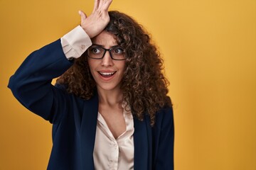 Hispanic woman with curly hair standing over yellow background surprised with hand on head for...