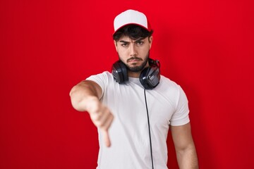 Hispanic man with beard wearing gamer hat and headphones looking unhappy and angry showing rejection and negative with thumbs down gesture. bad expression.