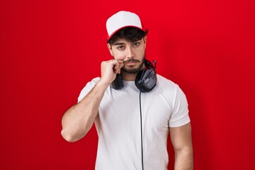 Hispanic man with beard wearing gamer hat and headphones mouth and lips shut as zip with fingers....