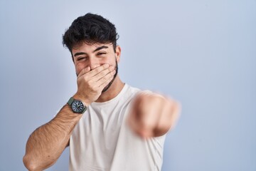 Hispanic man with beard standing over white background laughing at you, pointing finger to the camera with hand over mouth, shame expression