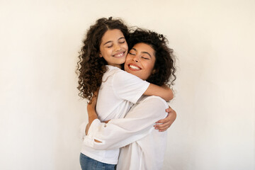 Joy of motherhood. Happy latin mother and daughter embracing, bonding and smiling over light wall...