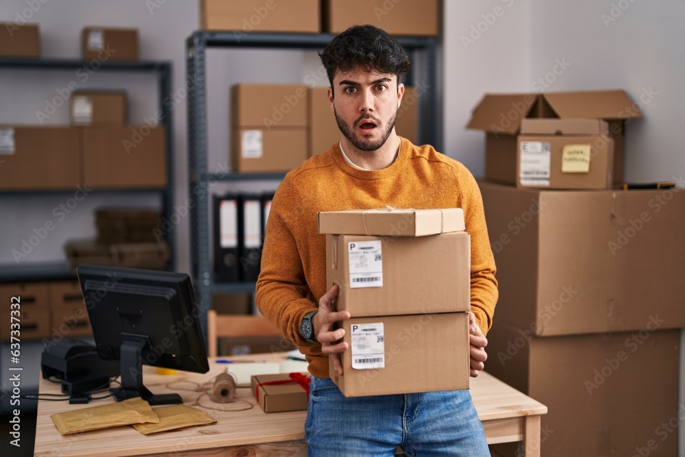 Wall mural Hispanic man with beard working at small business ecommerce holding packages in shock face, looking skeptical and sarcastic, surprised with open mouth