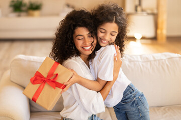 Mother's day concept. Adorable latin girl giving gift and embracing mom, mommy and daughter bonding together at home, sitting on sofa - obrazy, fototapety, plakaty