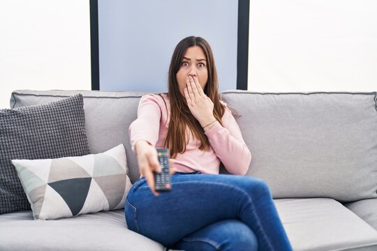 Young Brunette Woman Holding Television Remote Control Covering Mouth With Hand, Shocked And Afraid For Mistake. Surprised Expression