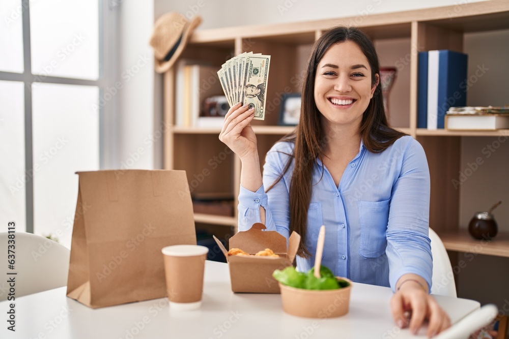 Sticker Young brunette woman eating take away food at home holding money looking positive and happy standing and smiling with a confident smile showing teeth