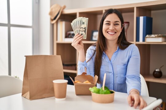 Young brunette woman eating take away food at home holding money looking positive and happy standing and smiling with a confident smile showing teeth