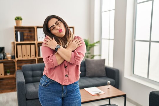 Young hispanic woman working at the office wearing glasses hugging oneself happy and positive, smiling confident. self love and self care