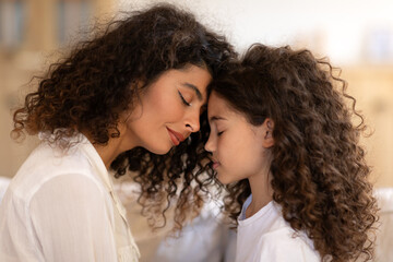 Tender moment. Loving mother and daughter bonding at home, touching each other with foreheads, side view. Happy latin mom and girl enjoying time together