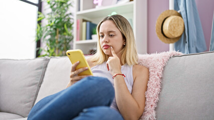 Young blonde woman using smartphone sitting on sofa thinking at home
