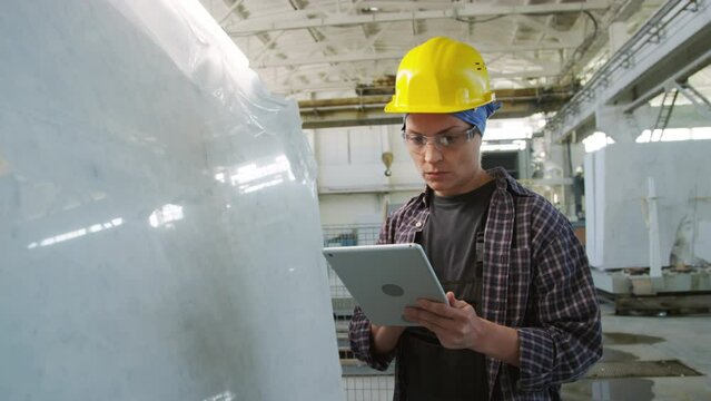 Medium shot of female quality control inspector in hardhat checking surface of white marble slab at stone factory and using digital tablet