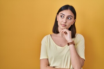 Hispanic girl wearing casual t shirt over yellow background with hand on chin thinking about question, pensive expression. smiling with thoughtful face. doubt concept.